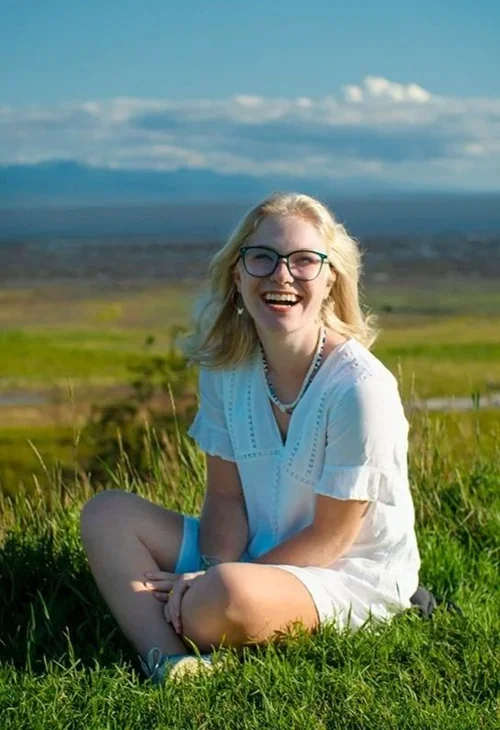 Smiling young woman with blonde hair and glasses sitting on green grass in a field, with a scenic landscape and blue sky in the background.