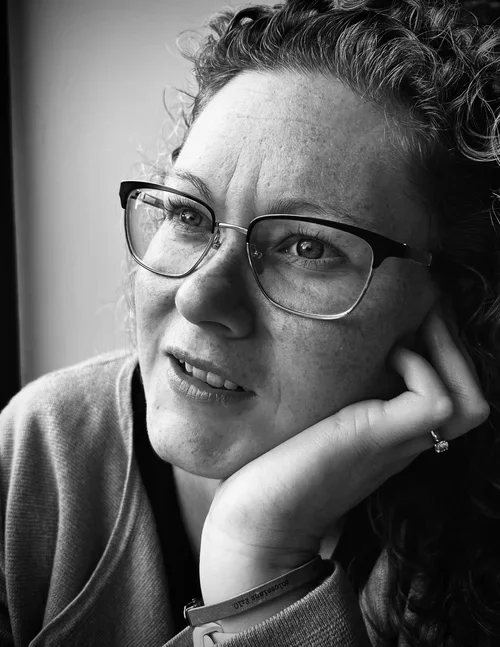 A woman with curly hair and glasses resting her chin on her hand, gazing thoughtfully outside the window in black and white.