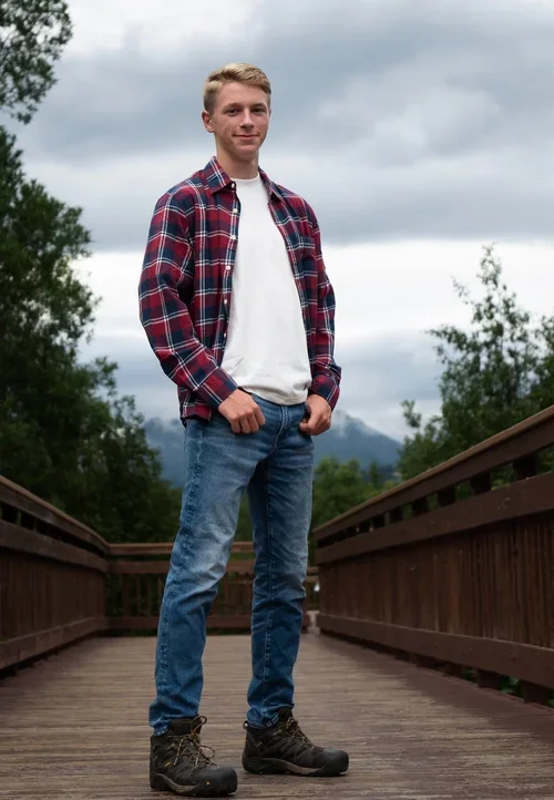 A young man standing on a wooden bridge outdoors, wearing a red plaid shirt, white t-shirt, jeans, and hiking boots, with trees and cloudy sky in the background.