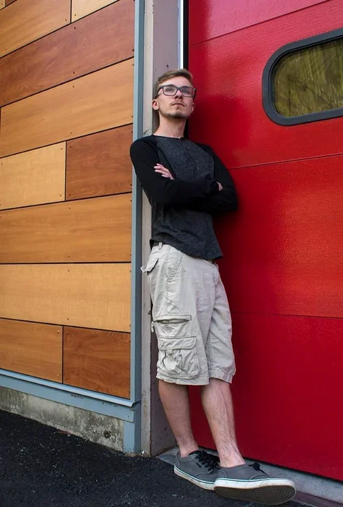 A young man with glasses and short hair standing against a red garage door and wooden wall, arms crossed, wearing a black and gray long-sleeve shirt, beige cargo shorts, and gray sneakers.