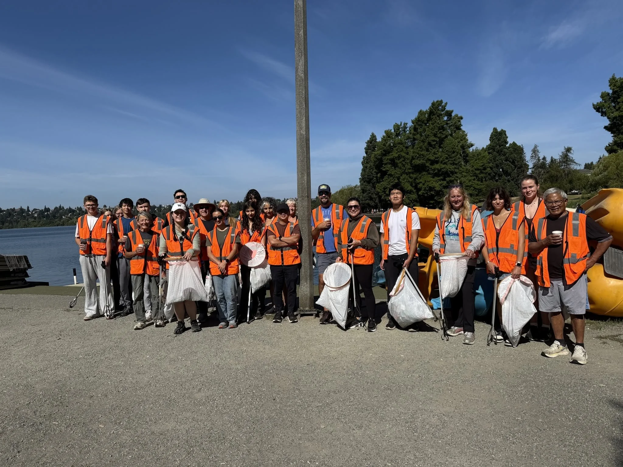 A group of people wearing orange safety vests and gathered outdoors near a body of water, with trees and a blue sky in the background, some holding white bags and umbrellas.