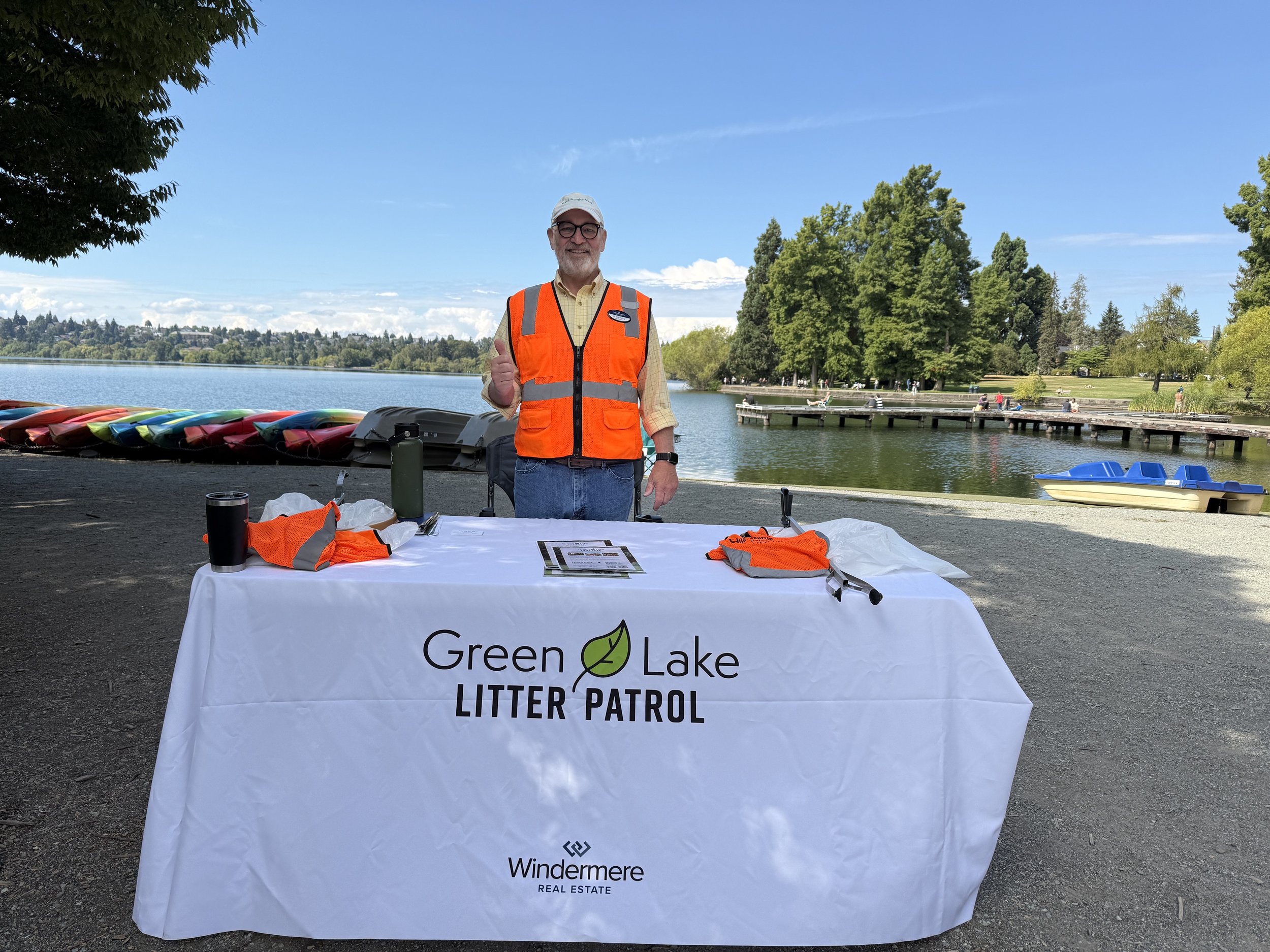 A man with glasses wearing an orange safety vest standing behind a table with a"Green Lake Litter Patrol" banner, near a lake with trees and people in the background.
