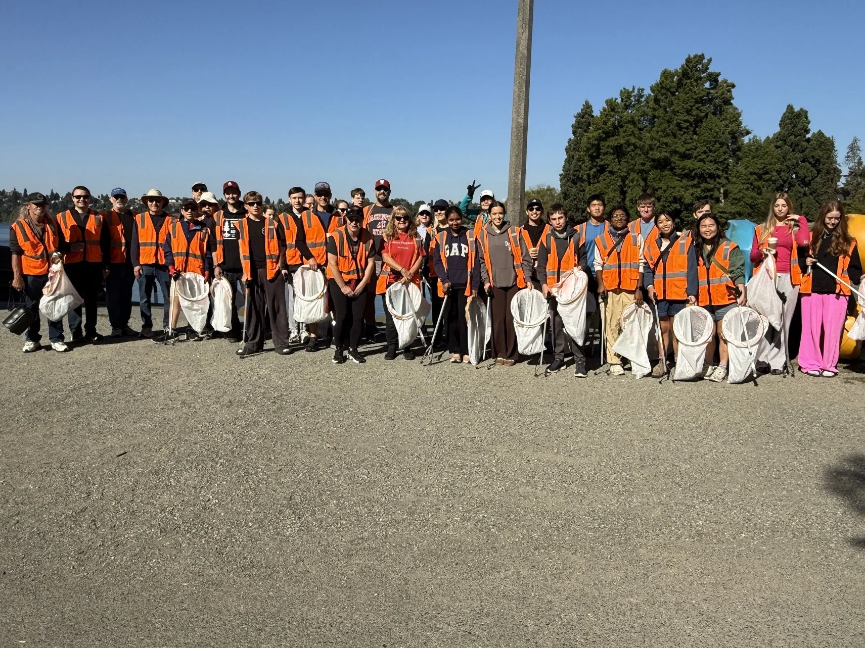 A large group of people wearing orange safety vests and holding collecting bags stands outdoors on a sunny day. They are smiling and posing for the photo with trees in the background.