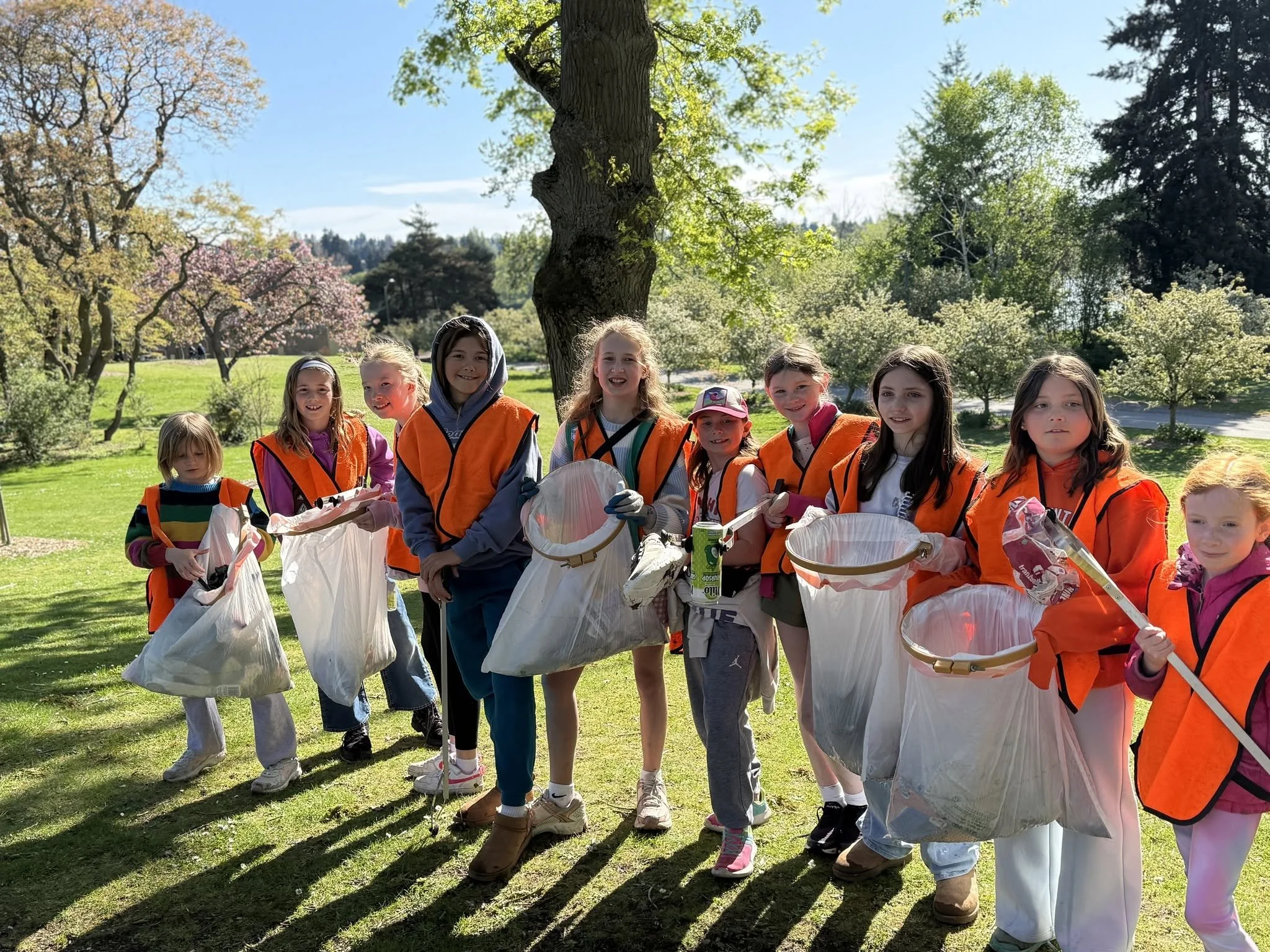 Group of children in orange vests holding trash bags during an outdoor cleanup event in a park on a sunny day.