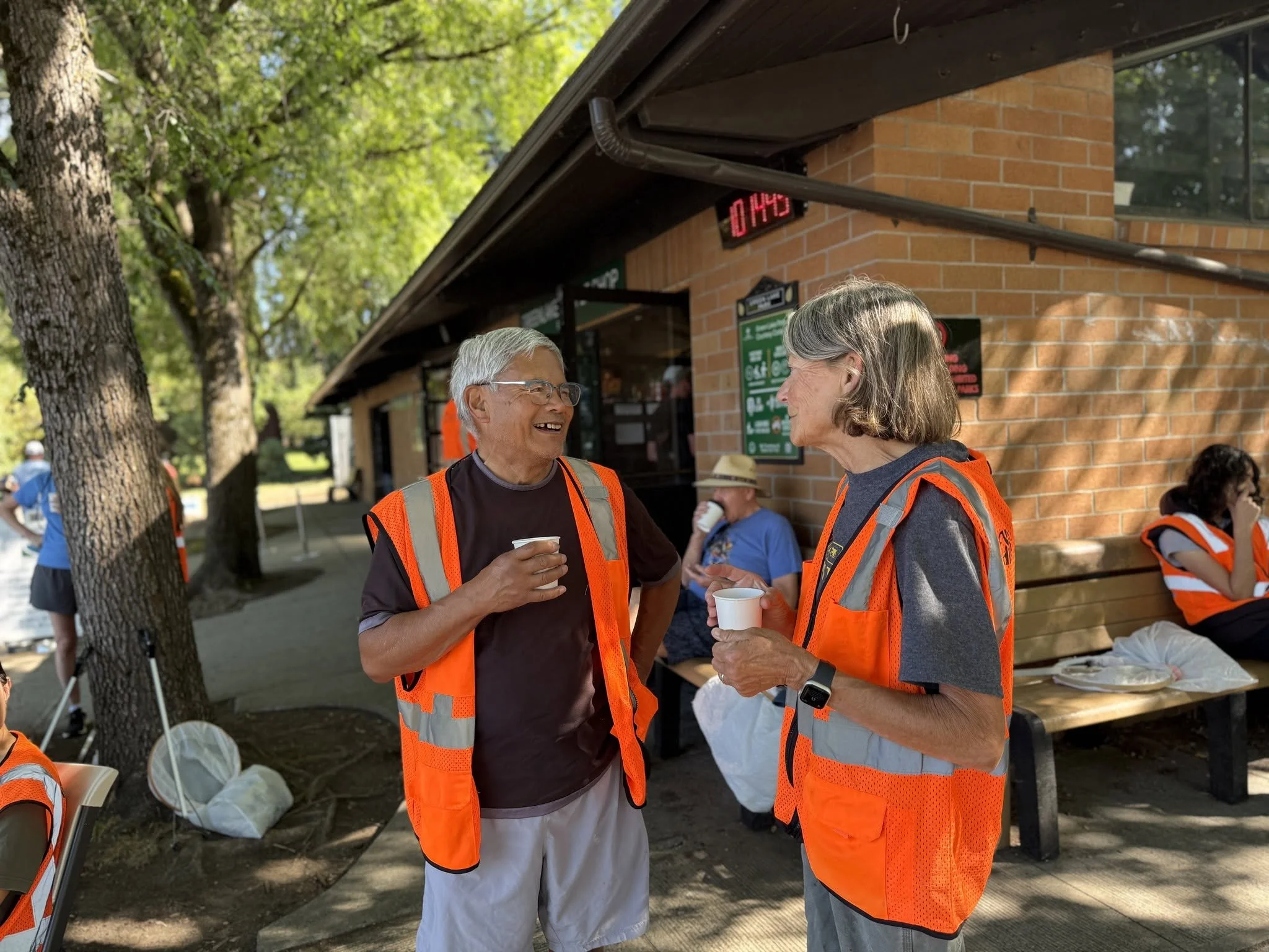 Two elderly people in orange safety vests talking and holding cups outdoors near a brick building, with trees in the background and others seated at tables.