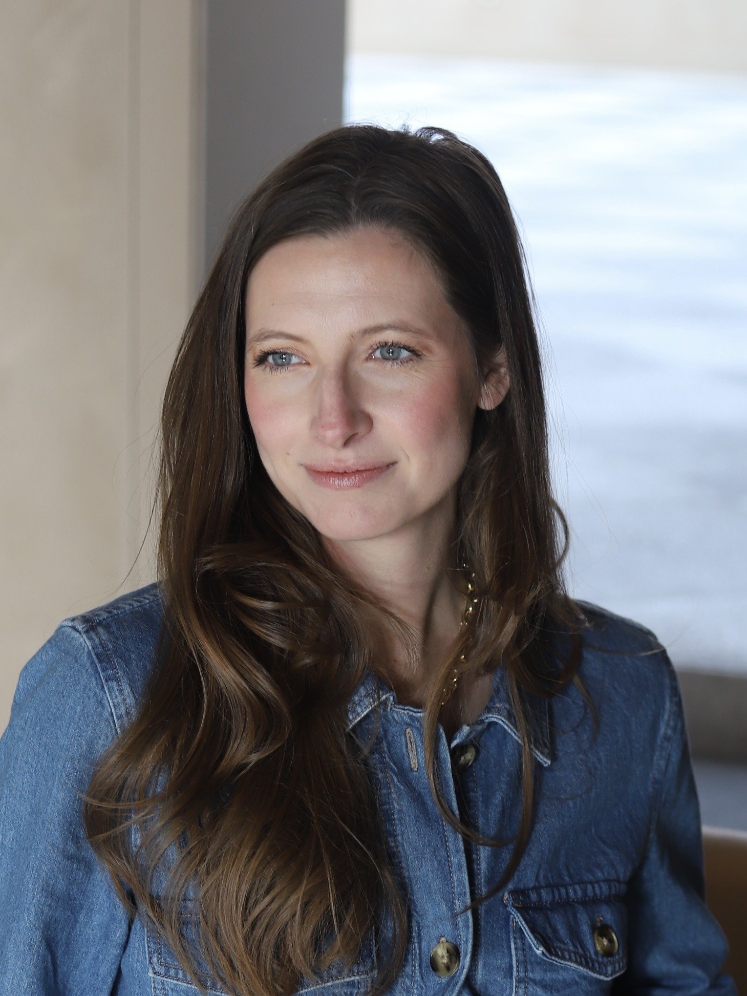 A woman with long wavy brown hair and blue eyes wearing a blue denim jacket, smiling gently, indoors near a window with daylight.