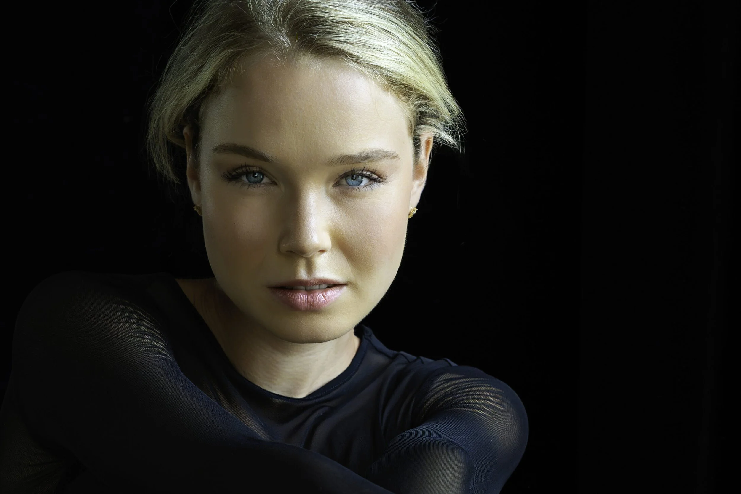 Nahaufnahme eines weiblichen Gesichts mit blauen Augen und kurzen blonden Haaren vor schwarzem Hintergrund.