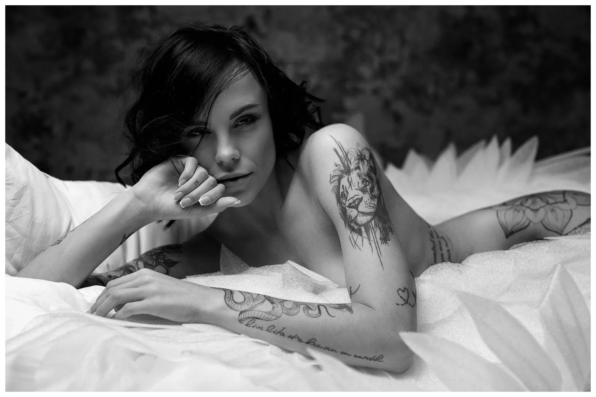 Schwarzer und weißer Foto einer jungen Frau mit Tattoos, die auf einem Bett liegt und in die Kamera schaut.