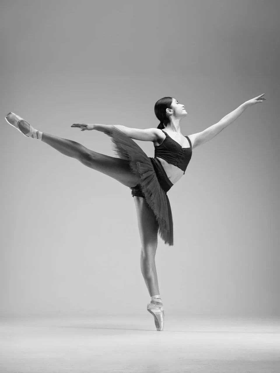 Eine Balletttänzerin in grauer Tutu und Spitzenschuhen macht eine Pirouette auf einem glatten Boden, schwarz-weißer Hintergrund.