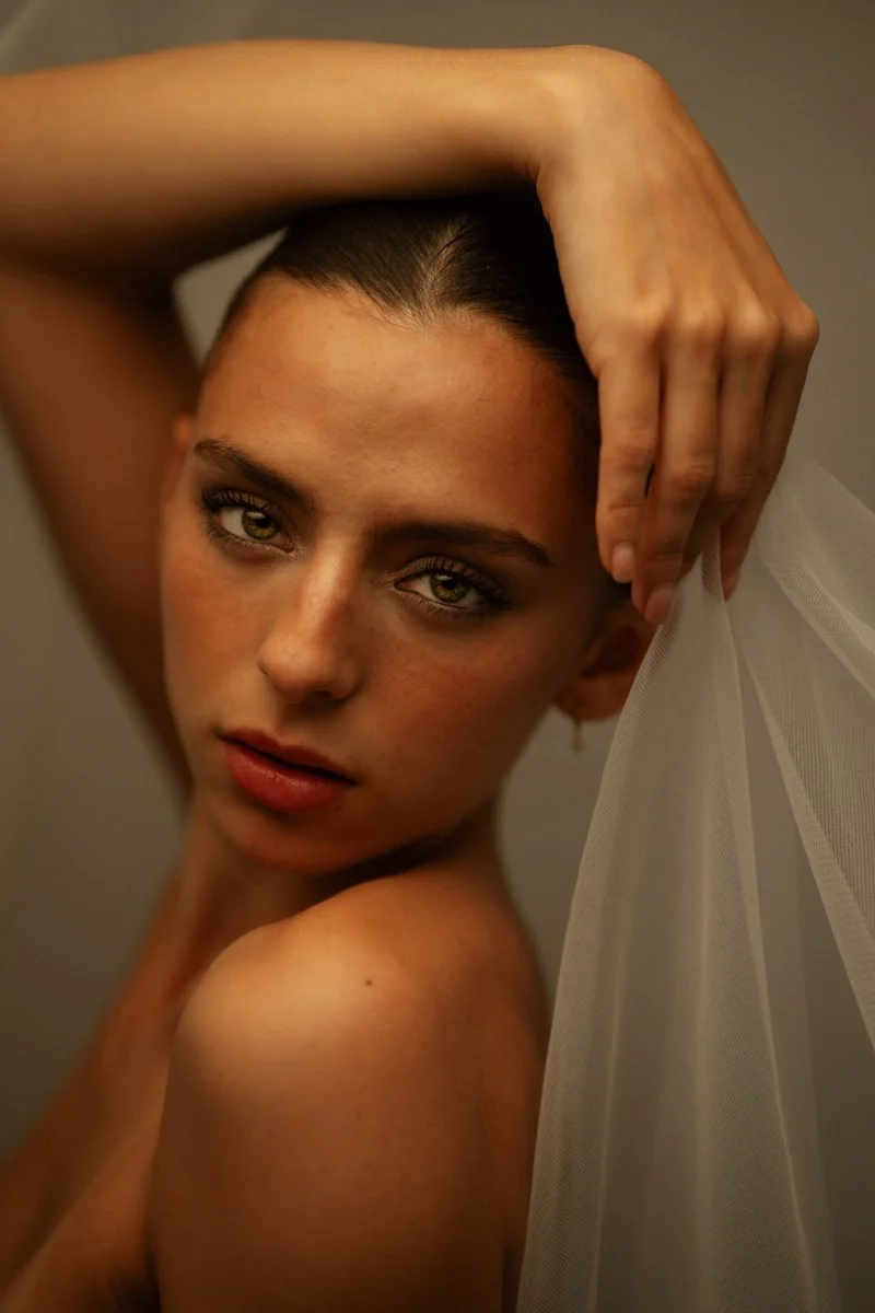 Nahaufnahme einer jungen Frau mit dunklem Haar, die sich an den Kopf fasst und an einer weißen Tüll- oder Organza-Textur lehnt.