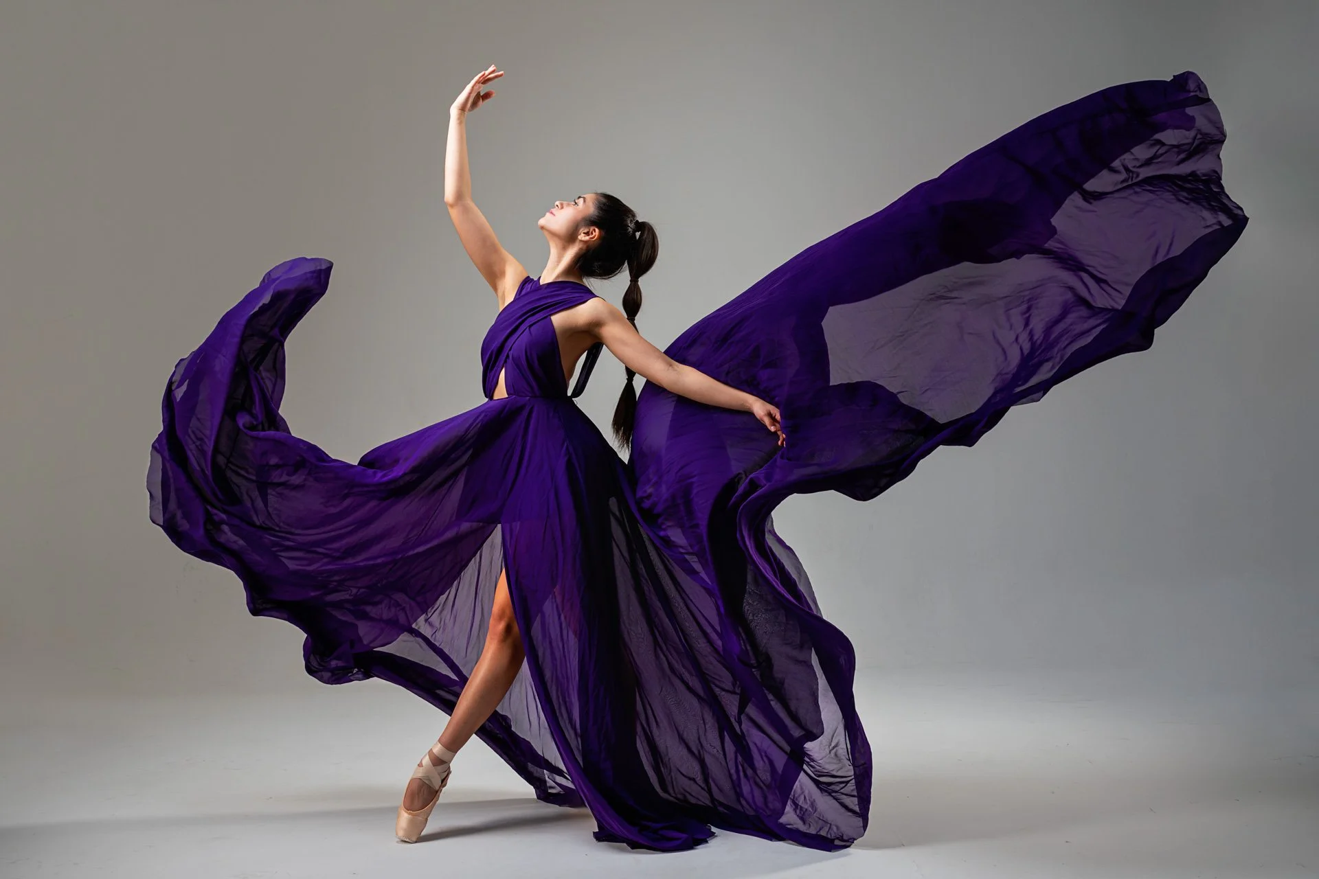 Balletttänzerin in einer langen, fließenden lila Robe, die sich im Wind bewegt, auf Zehenspitzen tanzend, mit grauem Hintergrund.