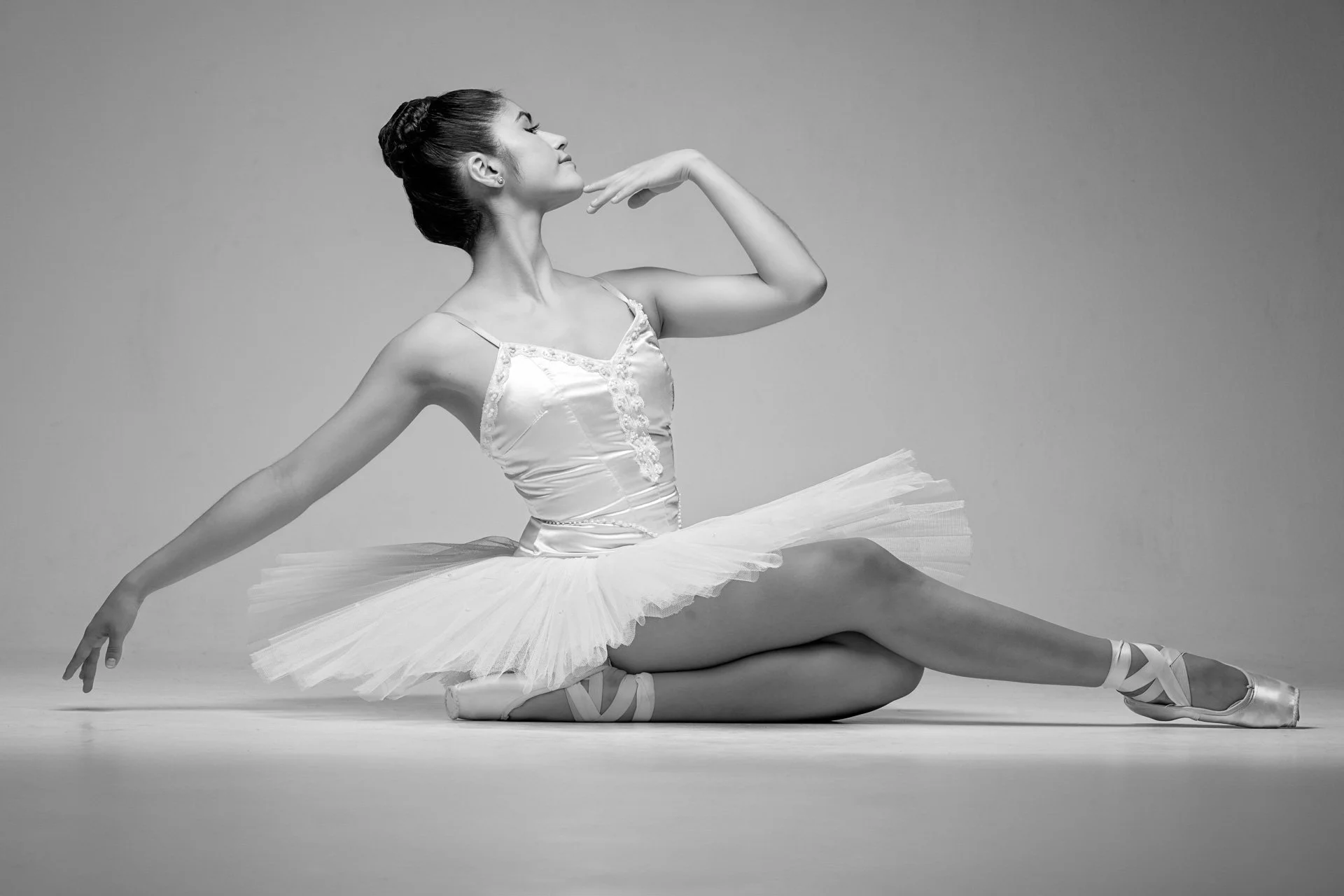 Elegante Balletttänzerin in weißem Kostüm, die eine Pose auf den Knien einnimmt, mit einem Tutu und Ballettschuhen, vor einem grauen Hintergrund.