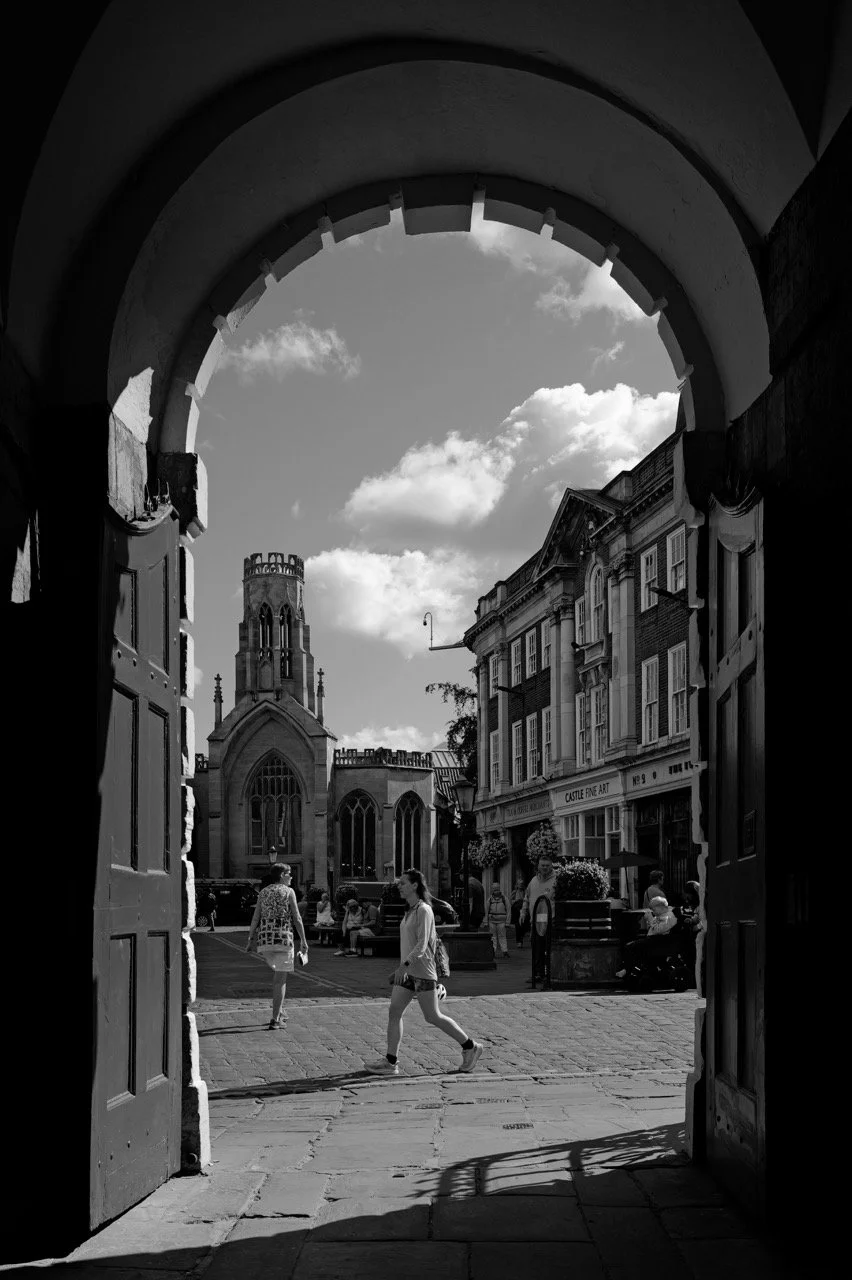 Blick durch eine offene Tür auf eine belebte Stadtstraße mit Menschen, historischen Gebäuden und einer Kirche im Hintergrund, bei sonnigem Himmel.