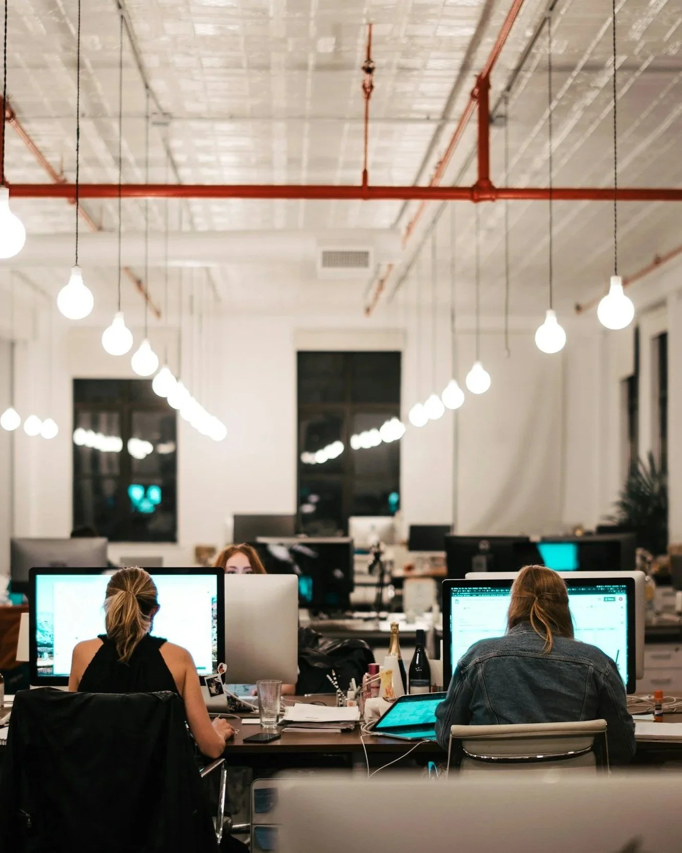 An open office space at night with multiple computer workstations, people working on computers, exposed ceiling with hanging light bulbs, and a large window in the background.