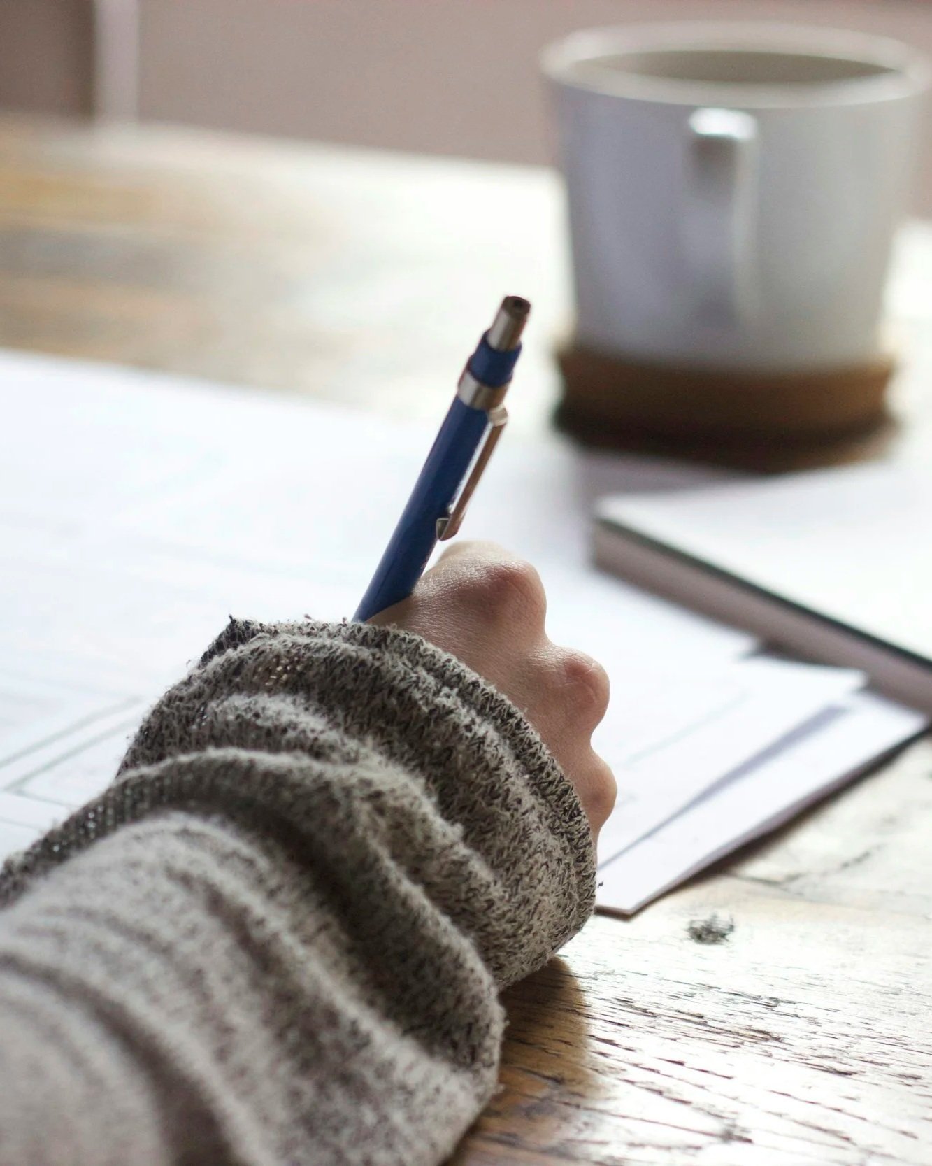 Close-up of a person's hand writing on a notebook with a blue pen, wearing a striped sweater, with a cup on a coaster in the background on a wooden table.
