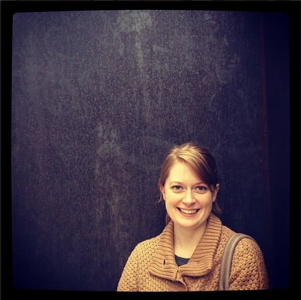 A woman with light brown hair smiling in front of a dark textured wall, wearing a brown knitted sweater and carrying a gray shoulder bag.