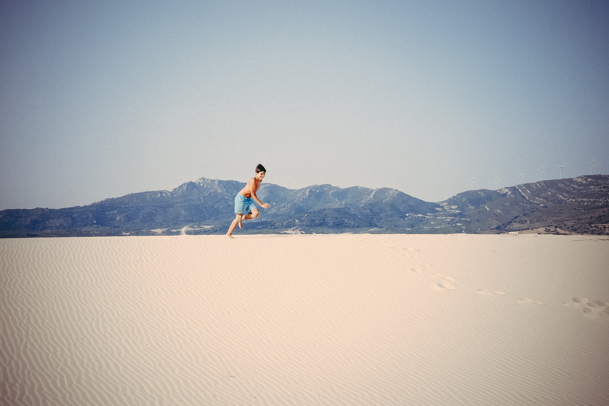 A young boy running or playing on sandy desert terrain with mountains in the background and wind turbines on the right side of the mountains.