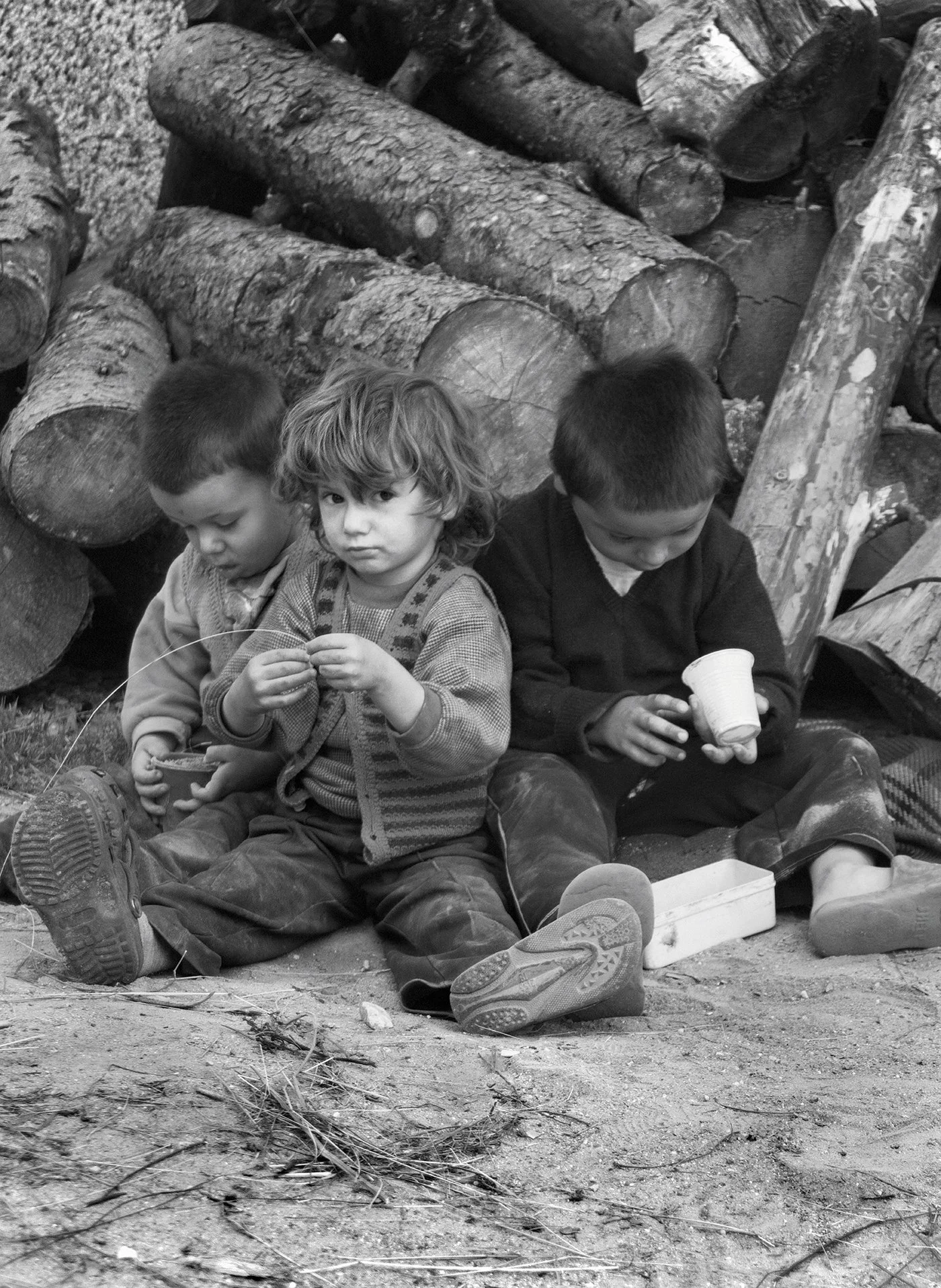 Three children sitting on the ground, surrounded by a pile of logs, with one of them holding a cup.