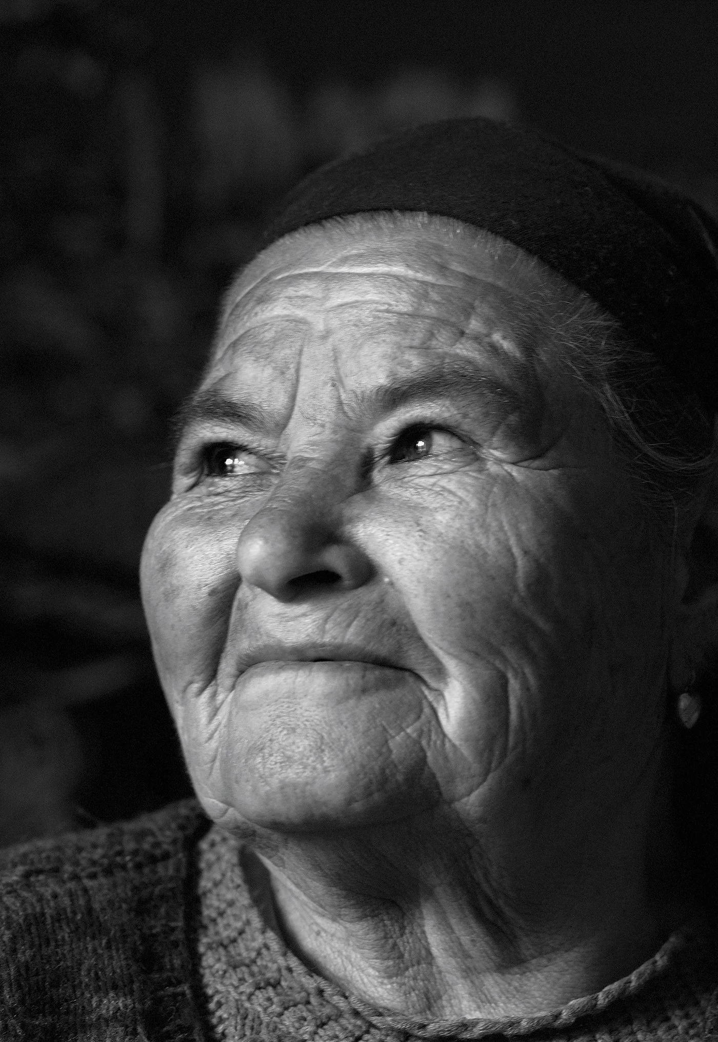 Close-up black and white photograph of an elderly woman's face, showing her eyes, wrinkles, and smile, with her head tilted upward.