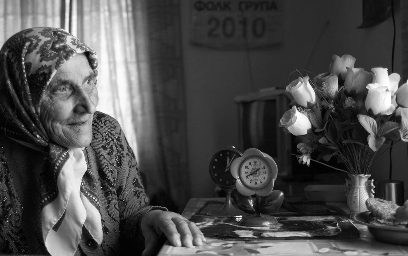 An elderly woman with a patterned headscarf sitting at a table with a vase of flowers, a clock, and a small food dish in a room with curtains and a calendar on the wall.
