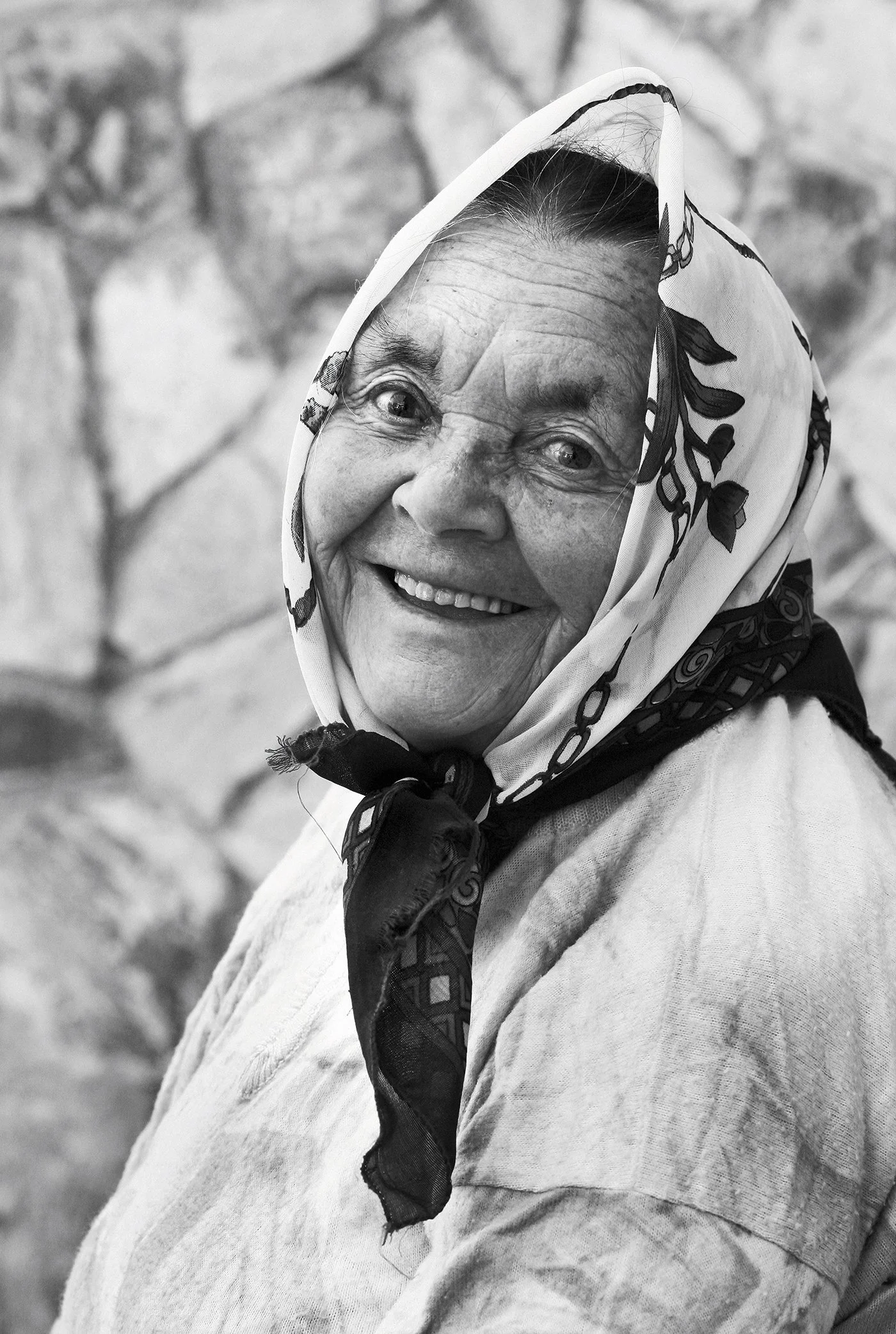 A smiling elderly woman wearing a headscarf with a floral pattern, looking directly at the camera against a blurred stone wall background.