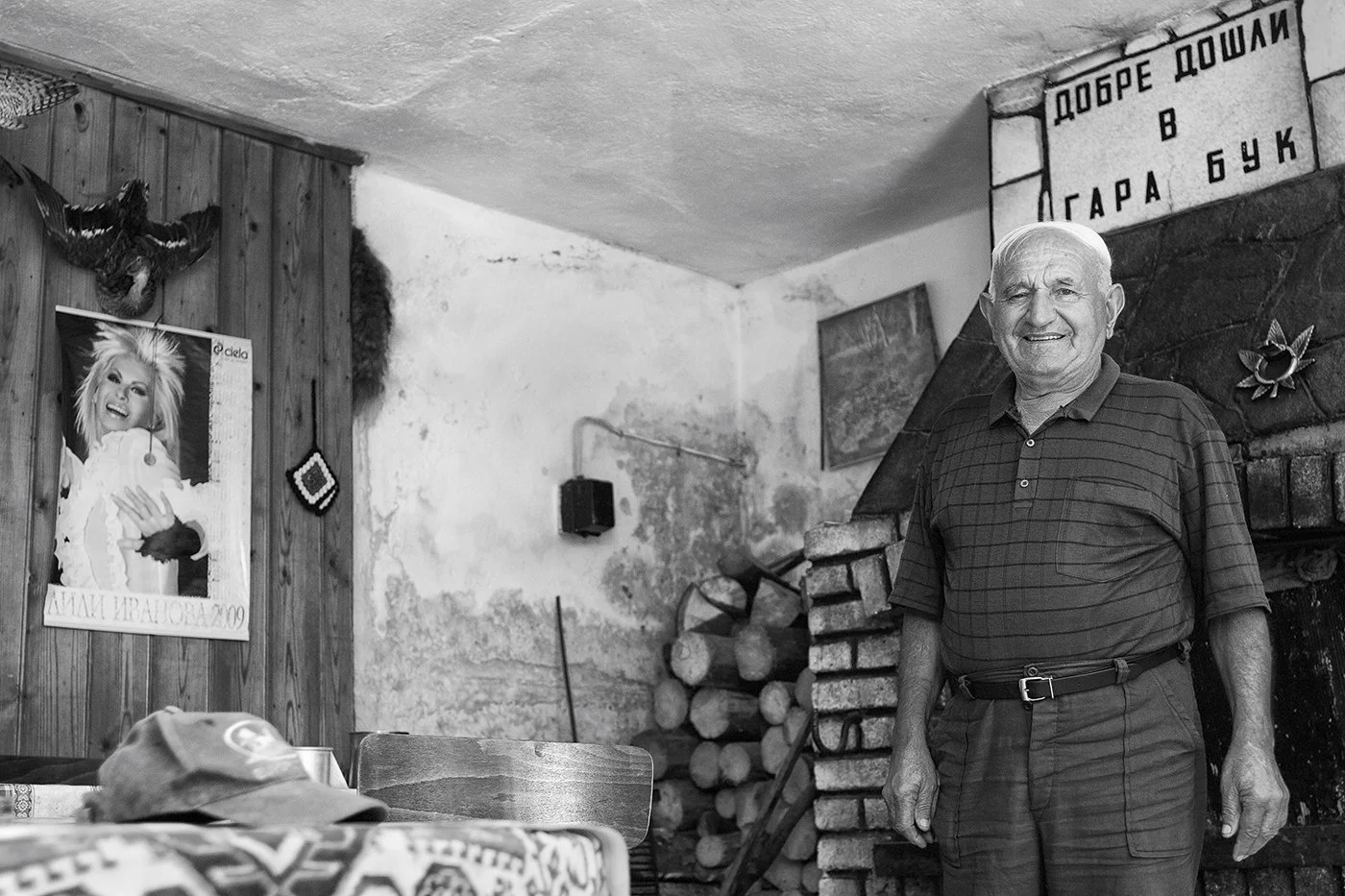 An elderly man standing inside a rustic room, smiling, with a stack of firewood and a fireplace behind him. The room has a poster on the wall and some decorations.