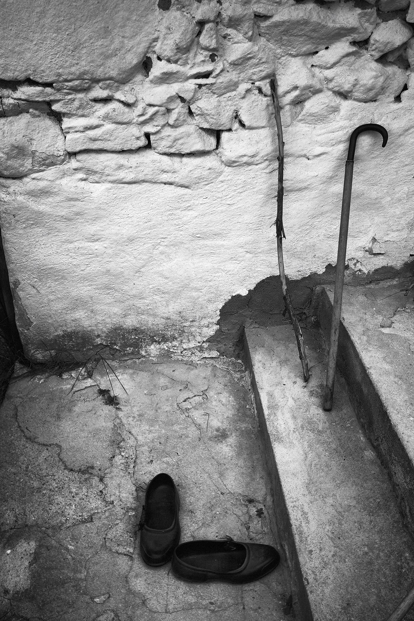 Black shoes on a cracked concrete floor with a rough stone wall in the background and a gardening fork leaning against the wall.