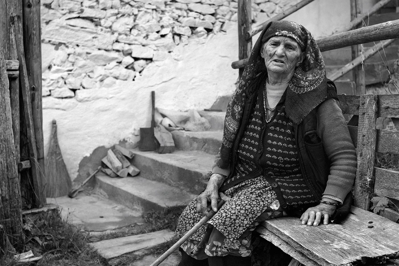 An elderly woman sitting outdoors on a wooden bench, wearing a patterned headscarf and layered clothing, holding a cane, with a rustic stone and wood structure in the background.
