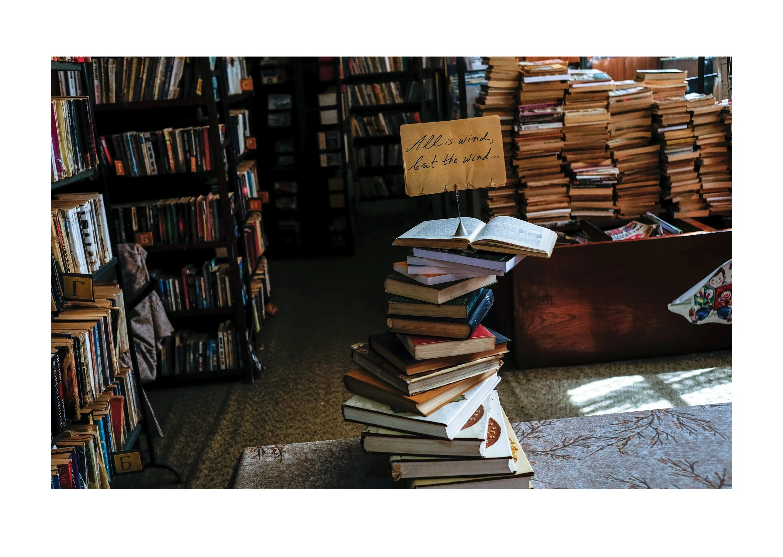 Library or bookstore with shelves filled with books and stacks of books on tables and chairs. A small handwritten sign reads 'All is wind, but the wind...'