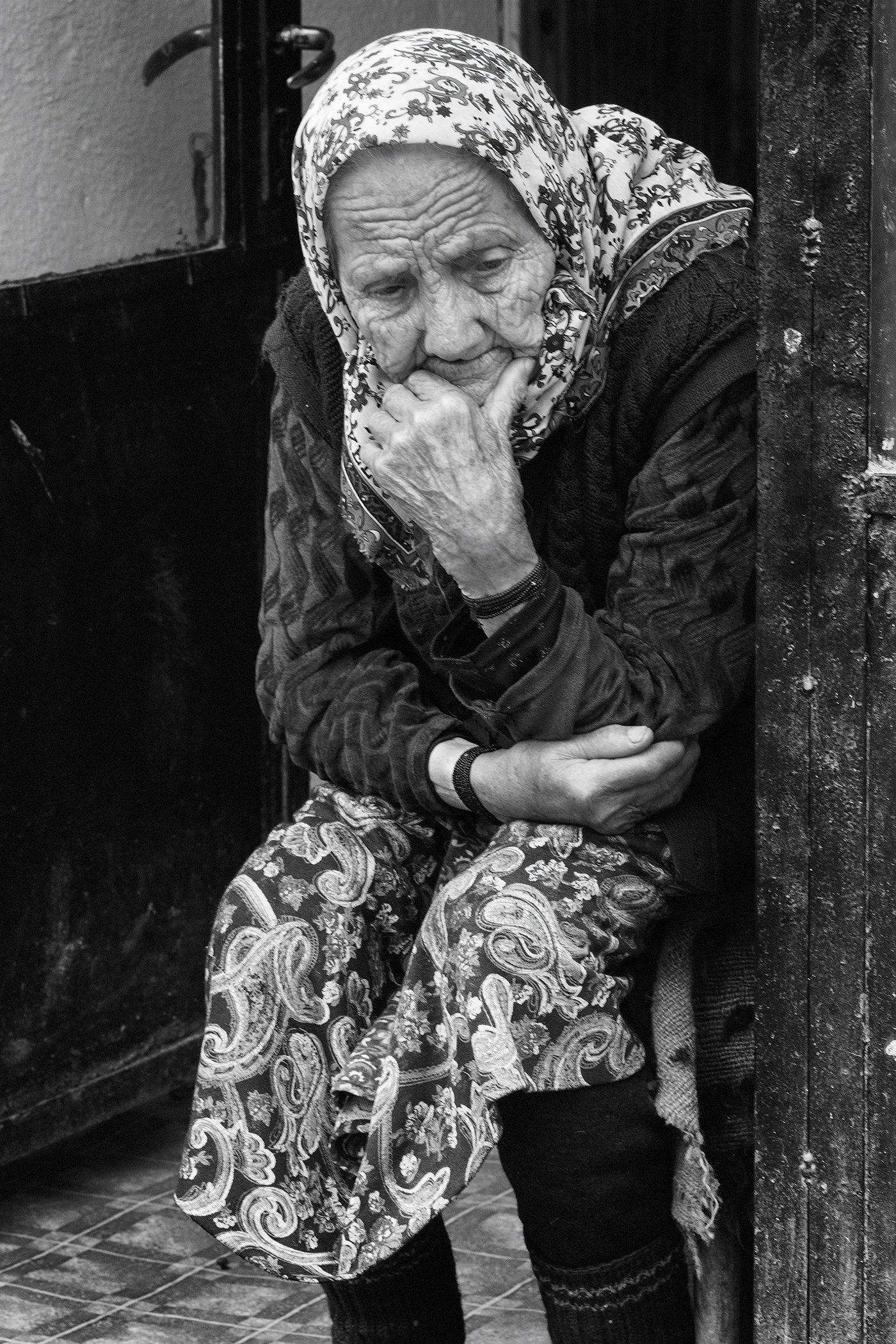 Black and white photo of an elderly woman with a patterned headscarf, sitting against a wall, with a thoughtful expression and hand on her chin.
