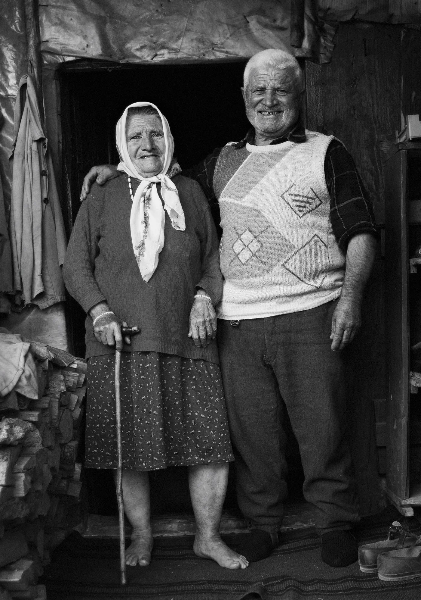 A black and white photo of an elderly woman and man standing close together indoors. The woman is wearing a headscarf, a sweater, and a skirt, holding a cane in her right hand. The man is wearing a patterned sweater over a collared shirt, with his right arm around the woman.