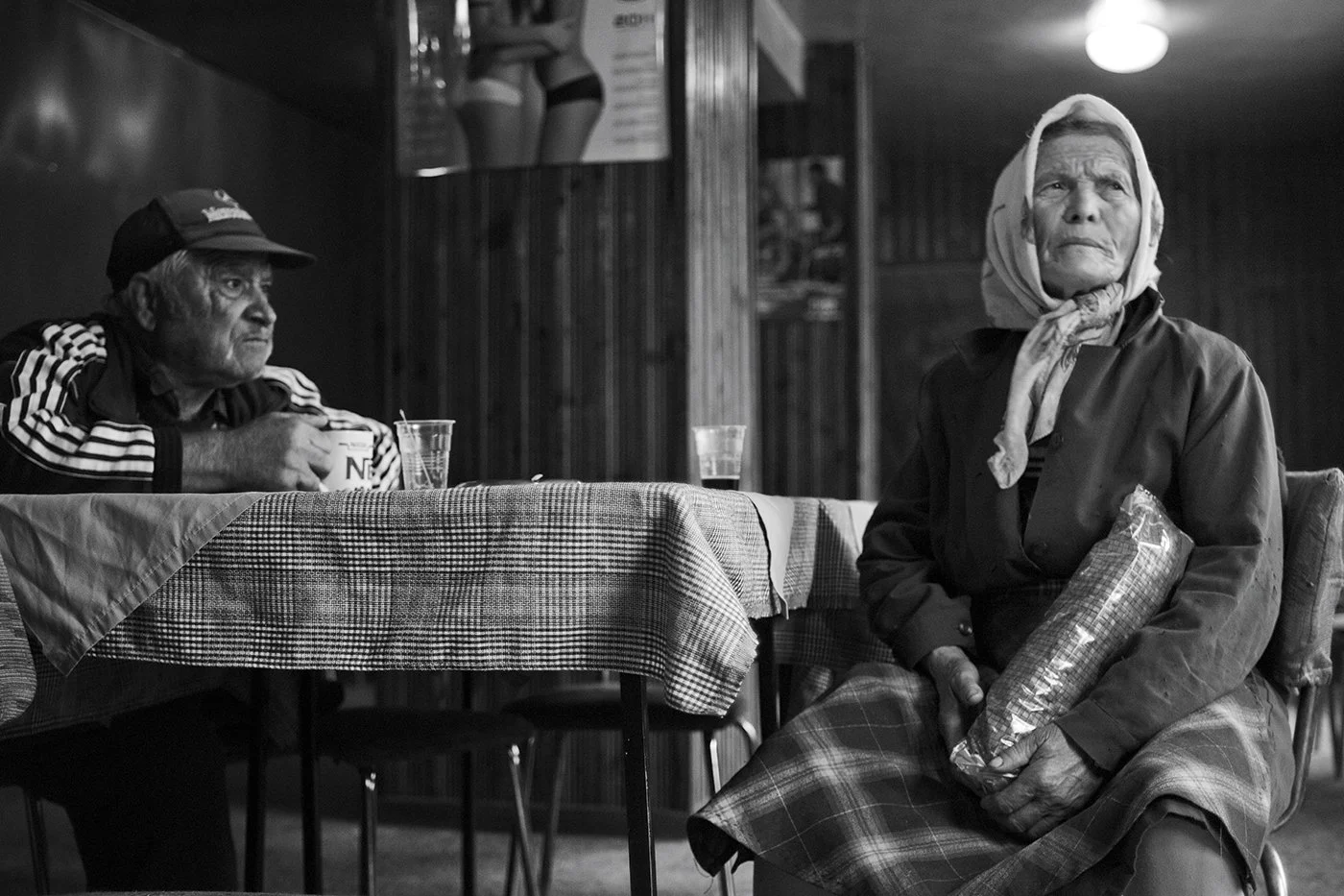 Two elderly individuals sitting at a table in a casual dining setting. The man on the left is wearing a cap and a track jacket, holding a mug, looking left with a serious expression. The woman on the right is wearing a headscarf and coat, holding a p