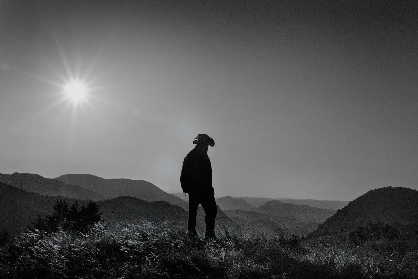 Silhouette of a person, possibly a cowboy, standing on a mountain ridge during sunset or sunrise, with rolling mountain ranges in the background and the sun shining brightly in the sky.