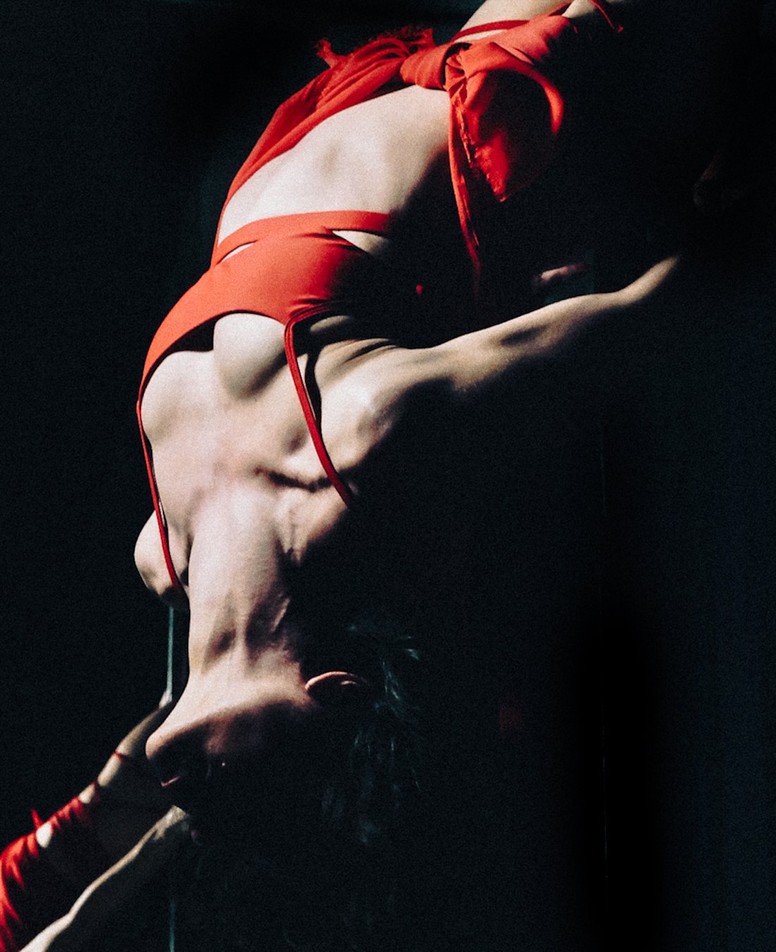 A woman in a red bikini and red ribbon highlights poses against a dark background, with her head tilted back and one arm bent, creating a dramatic silhouette.