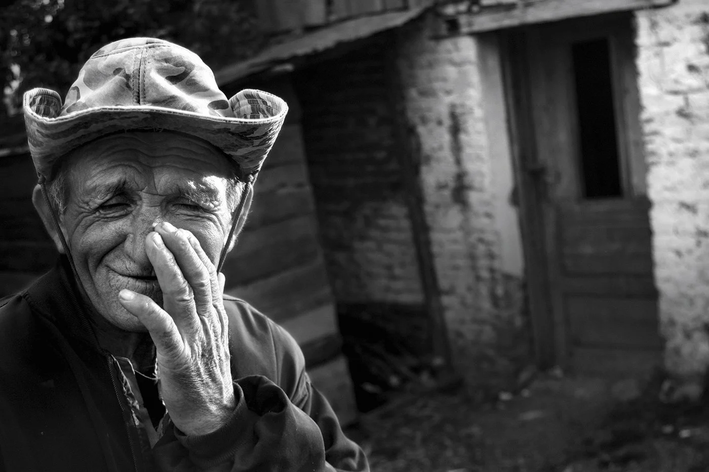 An elderly man wearing a camouflage hat and jacket, smiling and covering part of his face with his hand, standing outdoors near a rustic building.