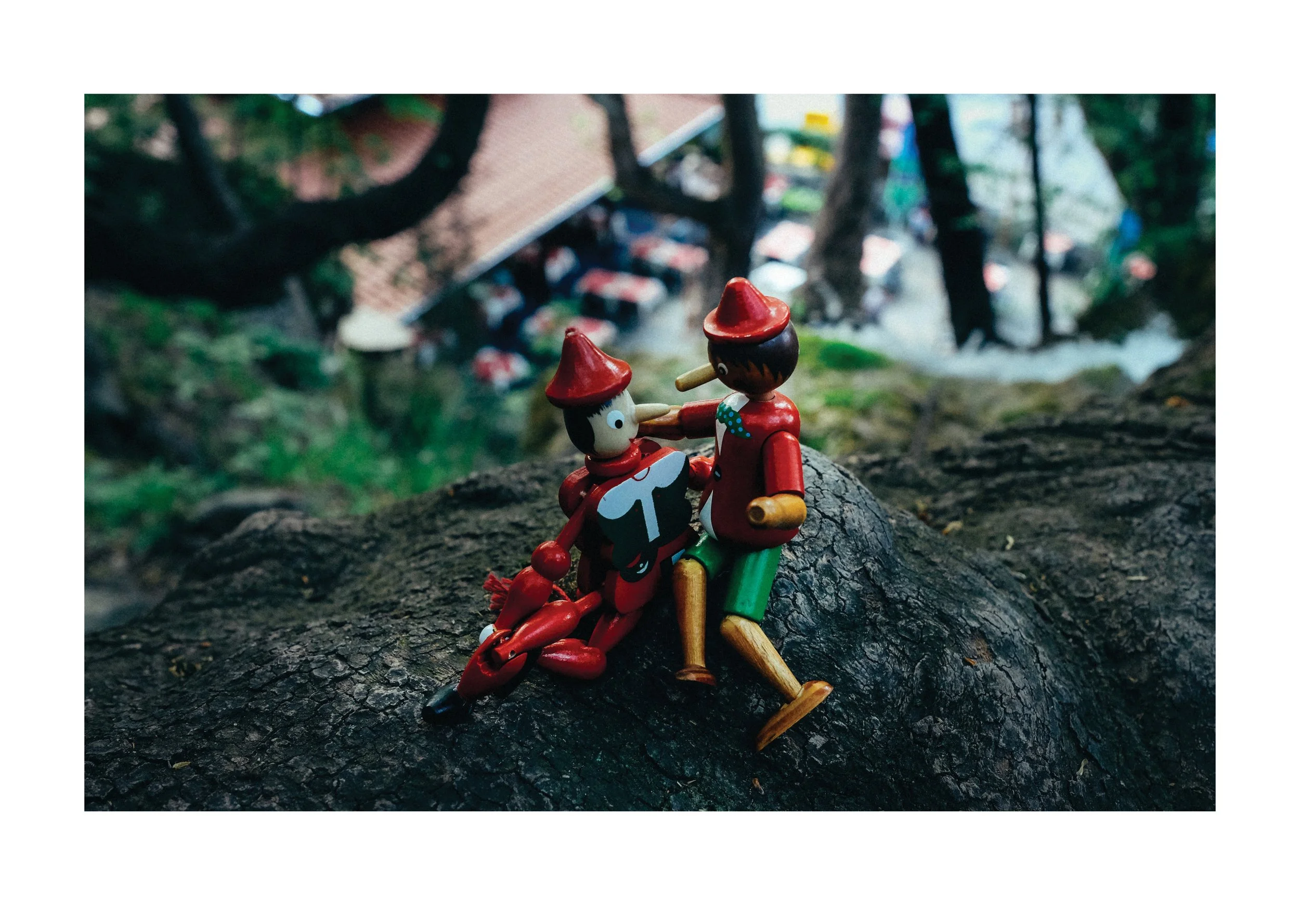 Wooden figurines of Pinocchio and a clown lying on a tree log in a wooded outdoor setting.