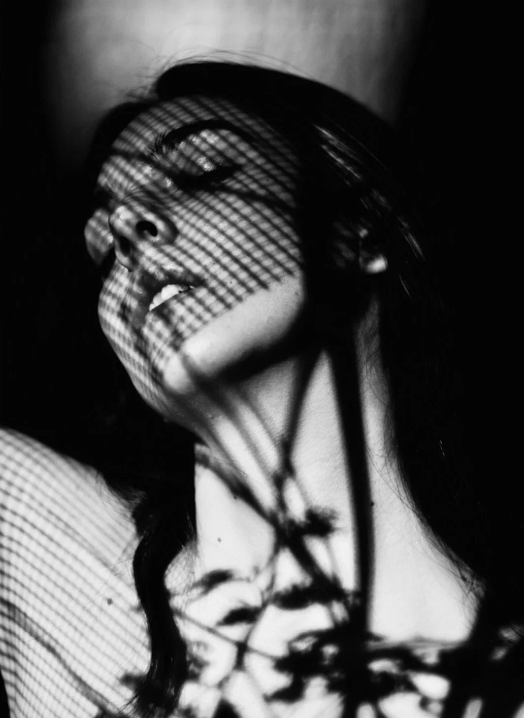 Black and white photo of a person with long hair with shadows of a textured grid cast across their face and hand.