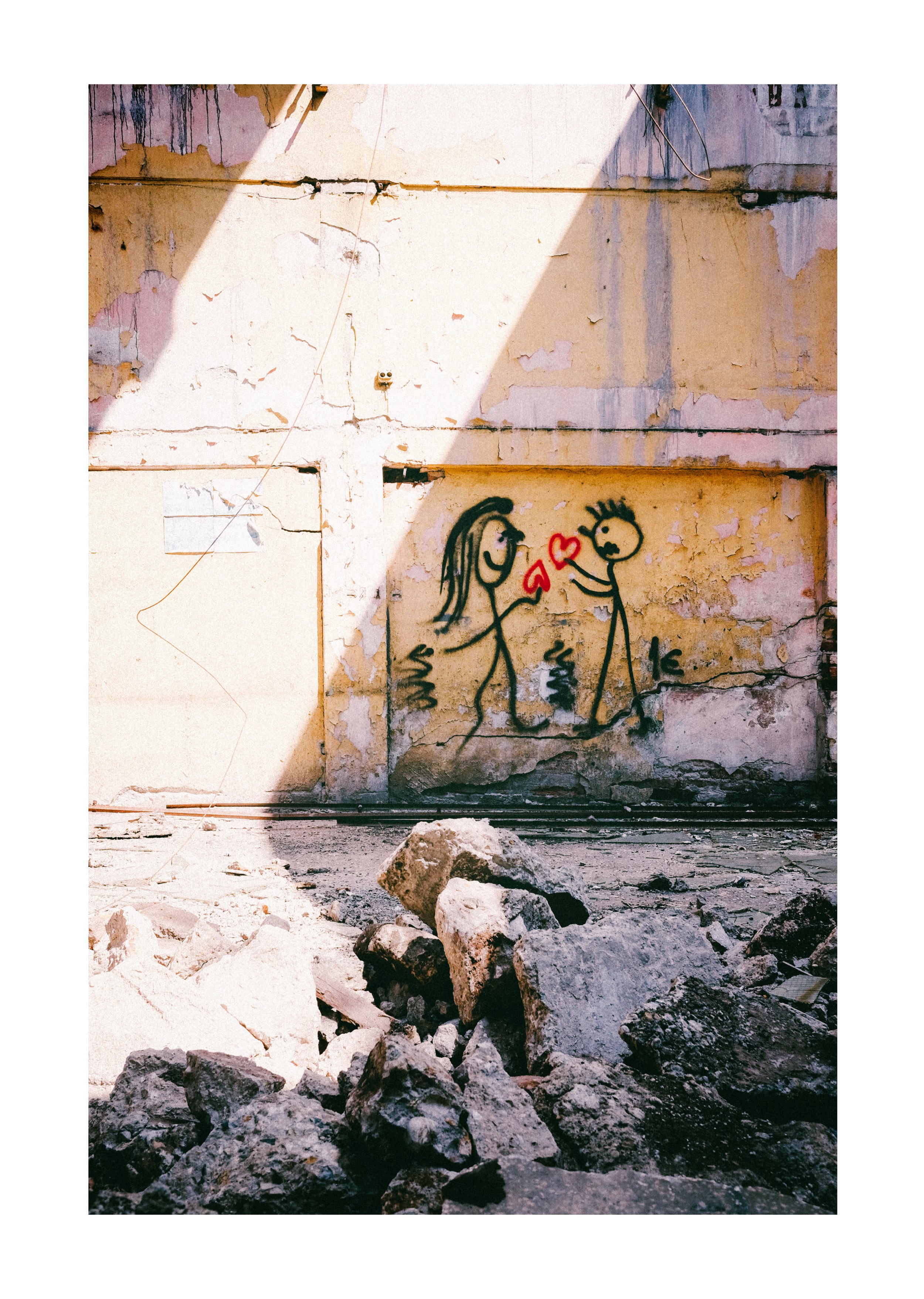 A wall with peeling paint, graffiti of two stick figures holding hearts, and rubble on the ground in front.