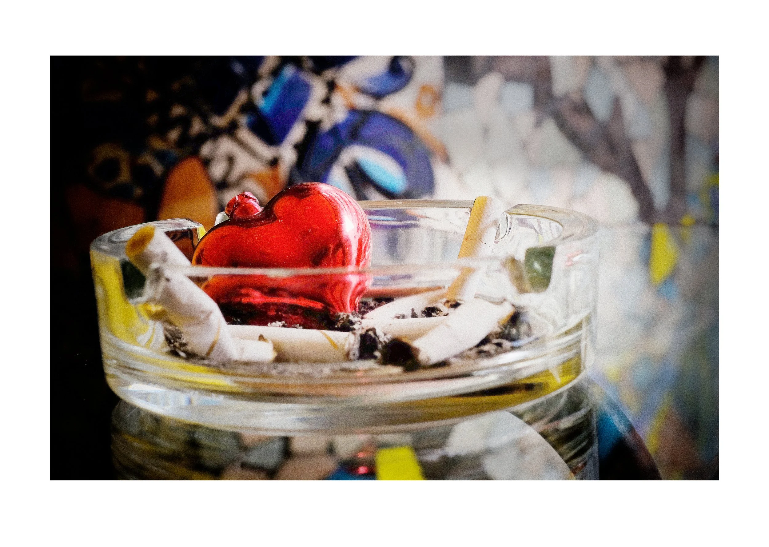 A glass bowl of a red and orange heart-shaped object, with some candies and crumpled wrapping paper inside, on a dark surface with a blurred colorful mosaic background.