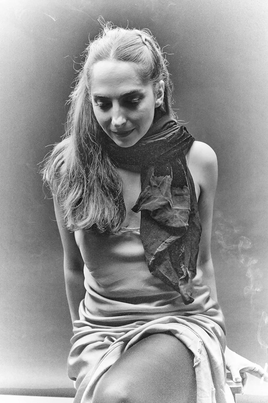 Black and white photo of a young woman sitting, with long hair, wearing a sleeveless top and a scarf around her neck, holding a cigarette in her right hand.