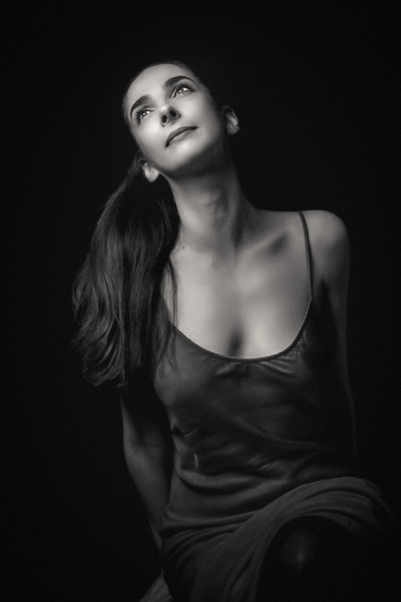 Black and white portrait of a woman with long hair wearing a dress with thin straps, looking upward with a thoughtful expression.