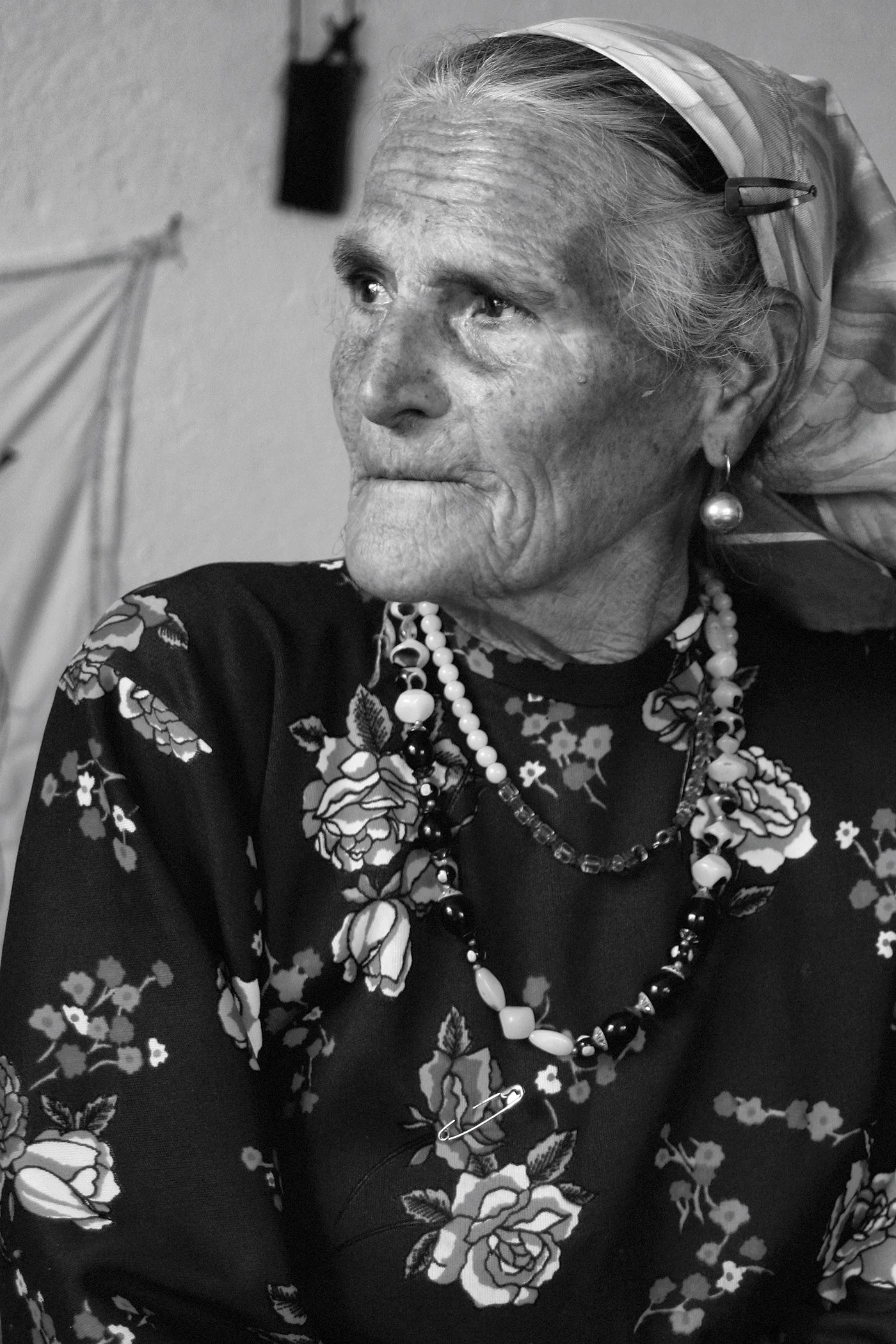 A black and white portrait of an elderly woman with light-colored hair pulled back, wearing large pearl earrings, a pearl necklace, and a floral patterned dark top. She gazes thoughtfully to her left.