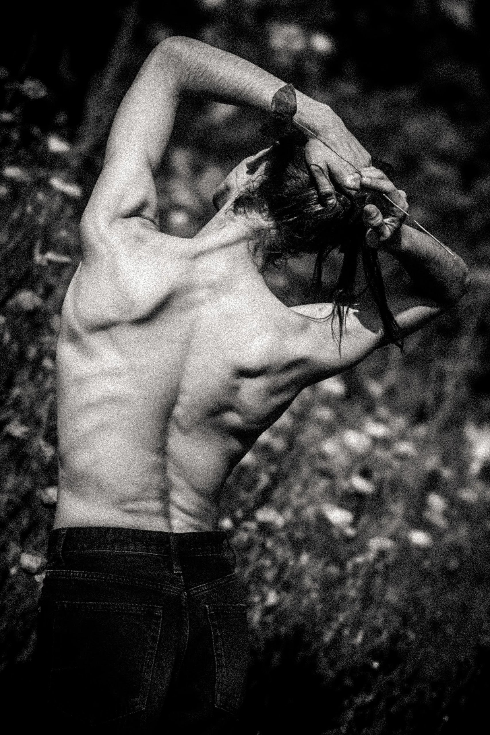 Black and white photo of a shirtless person with long hair, holding their head with one hand, standing outdoors with blurry foliage in the background.