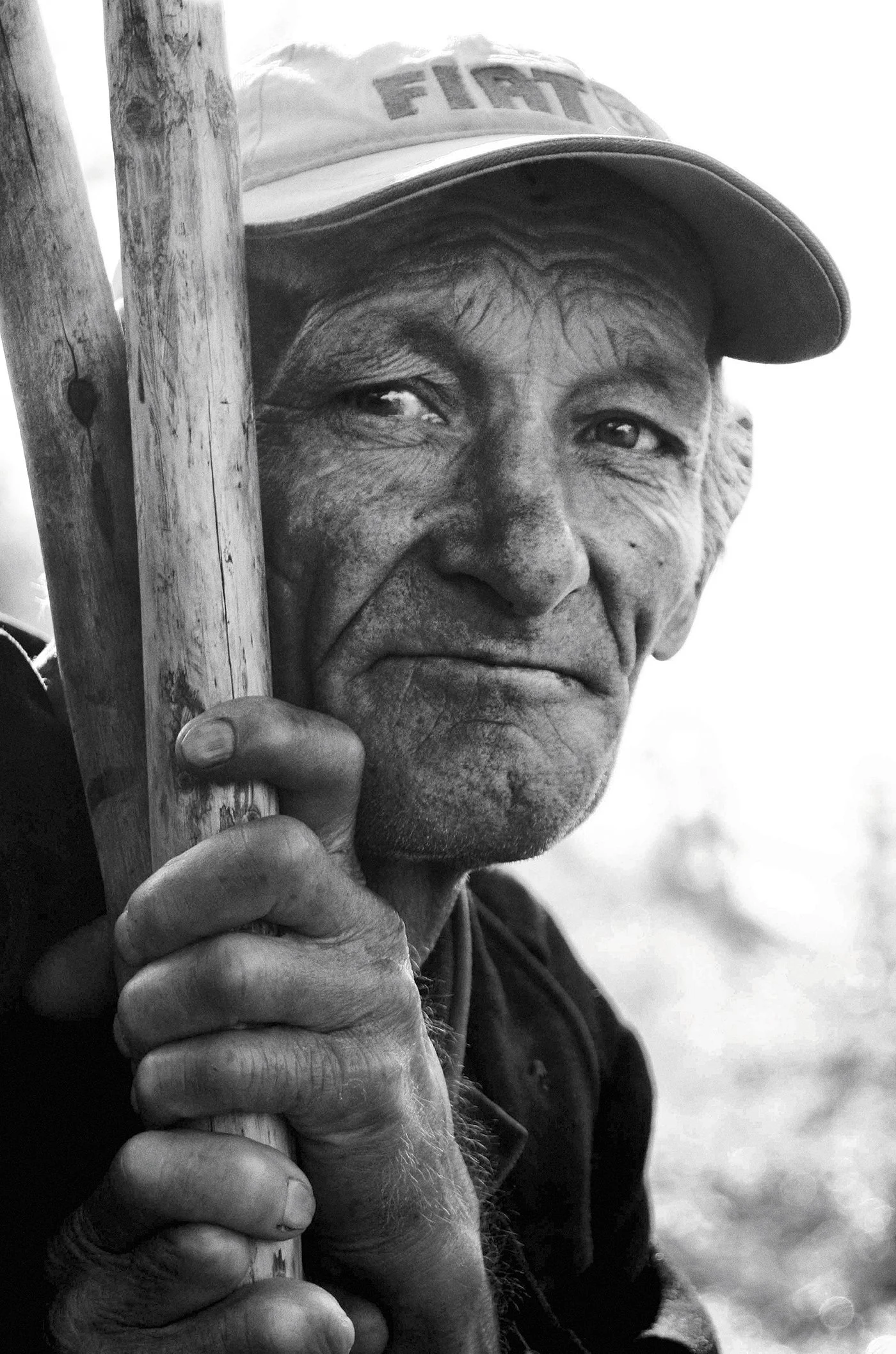 Close-up of an elderly man wearing a baseball cap with the text 'FIGHT' and holding a wooden stick close to his face, with a serious expression.
