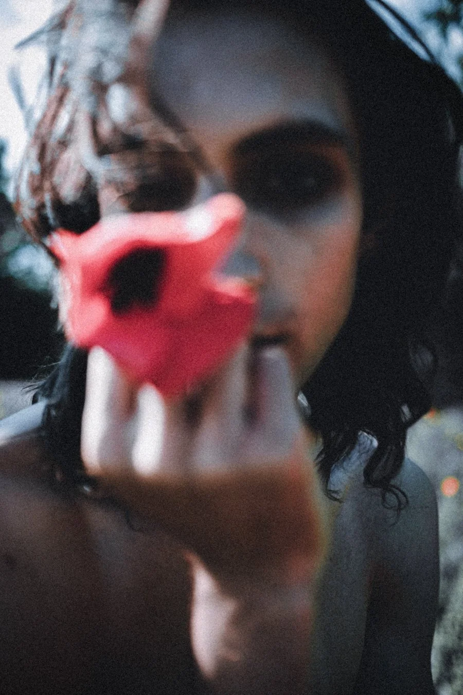 A woman with dark curly hair and dark makeup holding a small red object close to the camera, partially obscuring her face, with a blurred outdoor background.