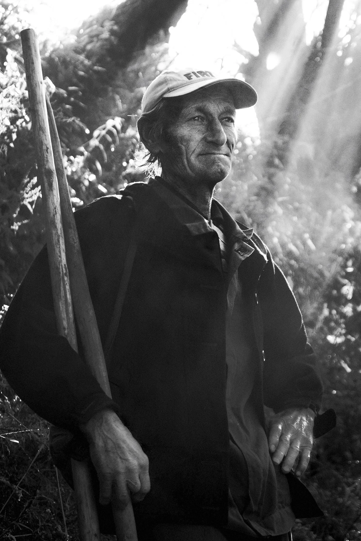 A black and white photo of an older man with weathered skin wearing a baseball cap and outdoors, holding a wooden walking stick, surrounded by trees and natural sunlight.