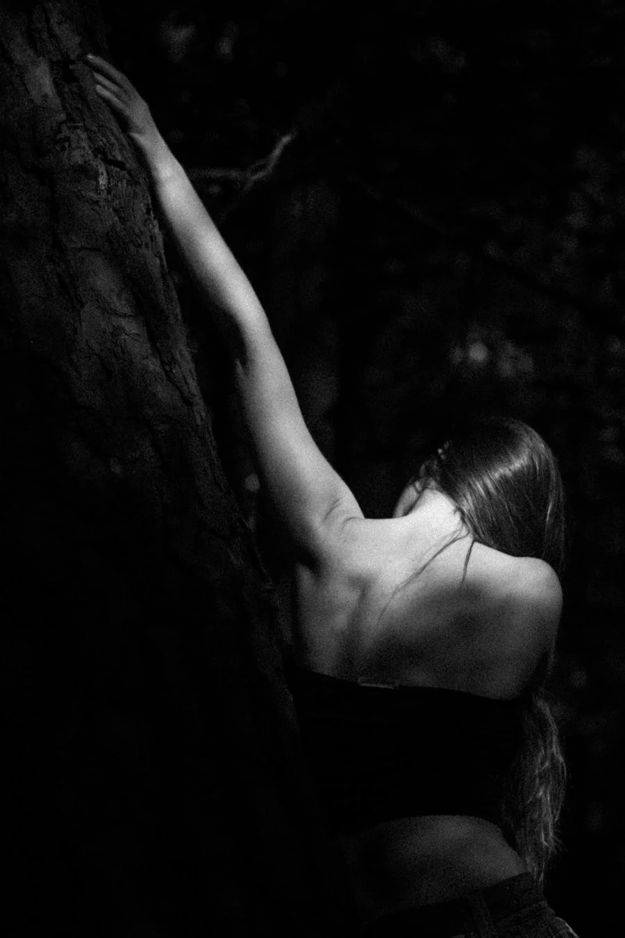 A person with long hair leaning against a tree at night with their arm extended up the tree.