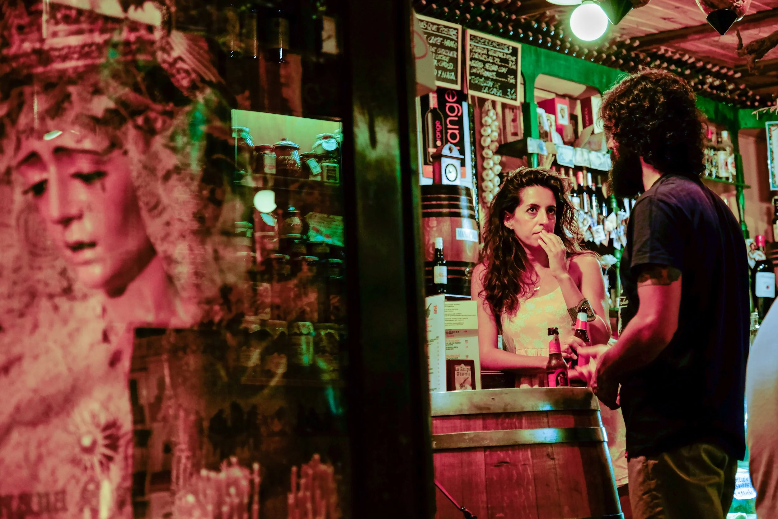 Two women are talking at a bar with colorful lighting. One woman has curly hair and is holding a bottle, the other woman has straight hair and appears to be listening.