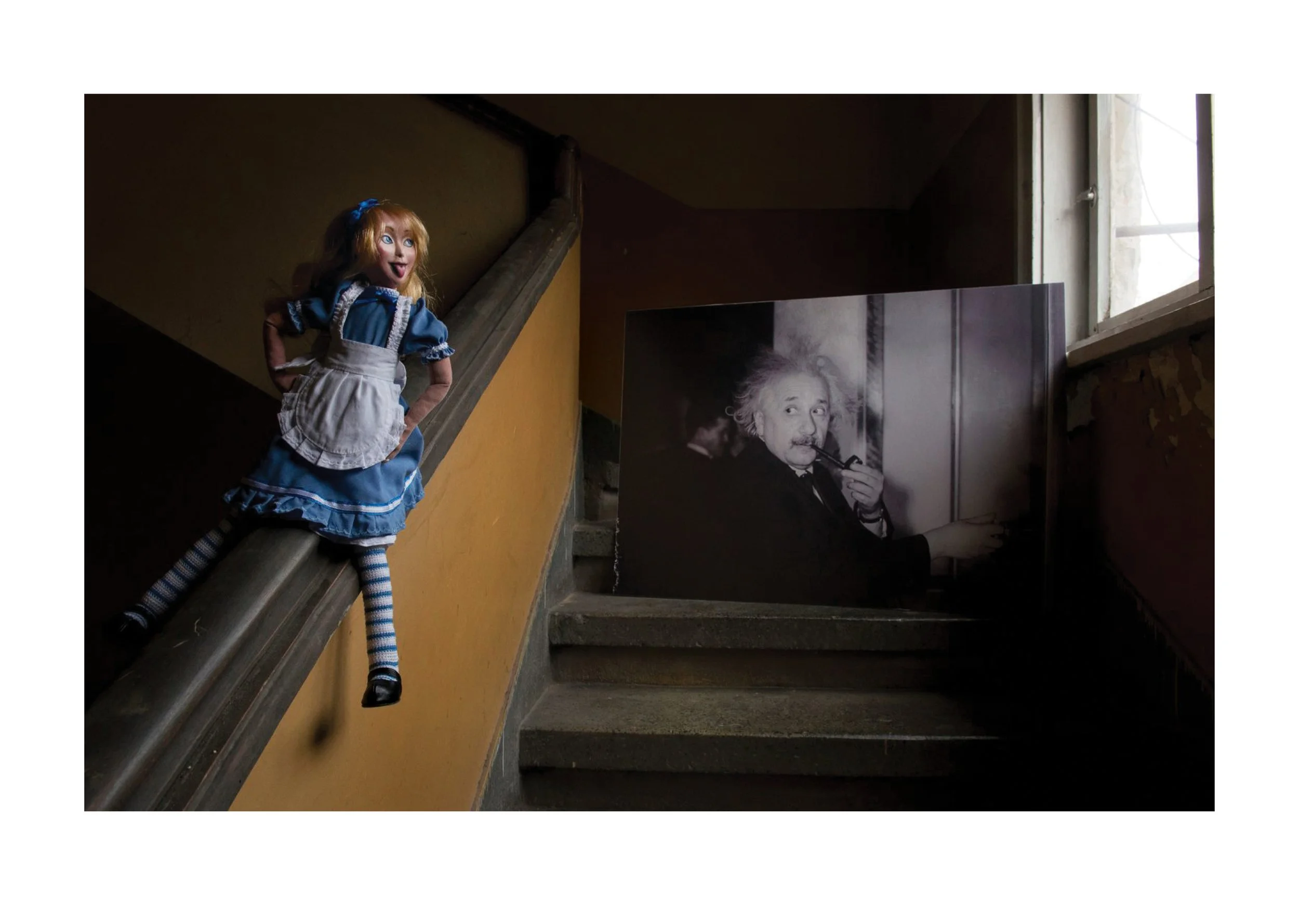 A doll with red hair, blue eyes, and a blue dress with a white apron, sitting on a staircase next to a black and white photograph of Albert Einstein, in a dimly lit interior space.