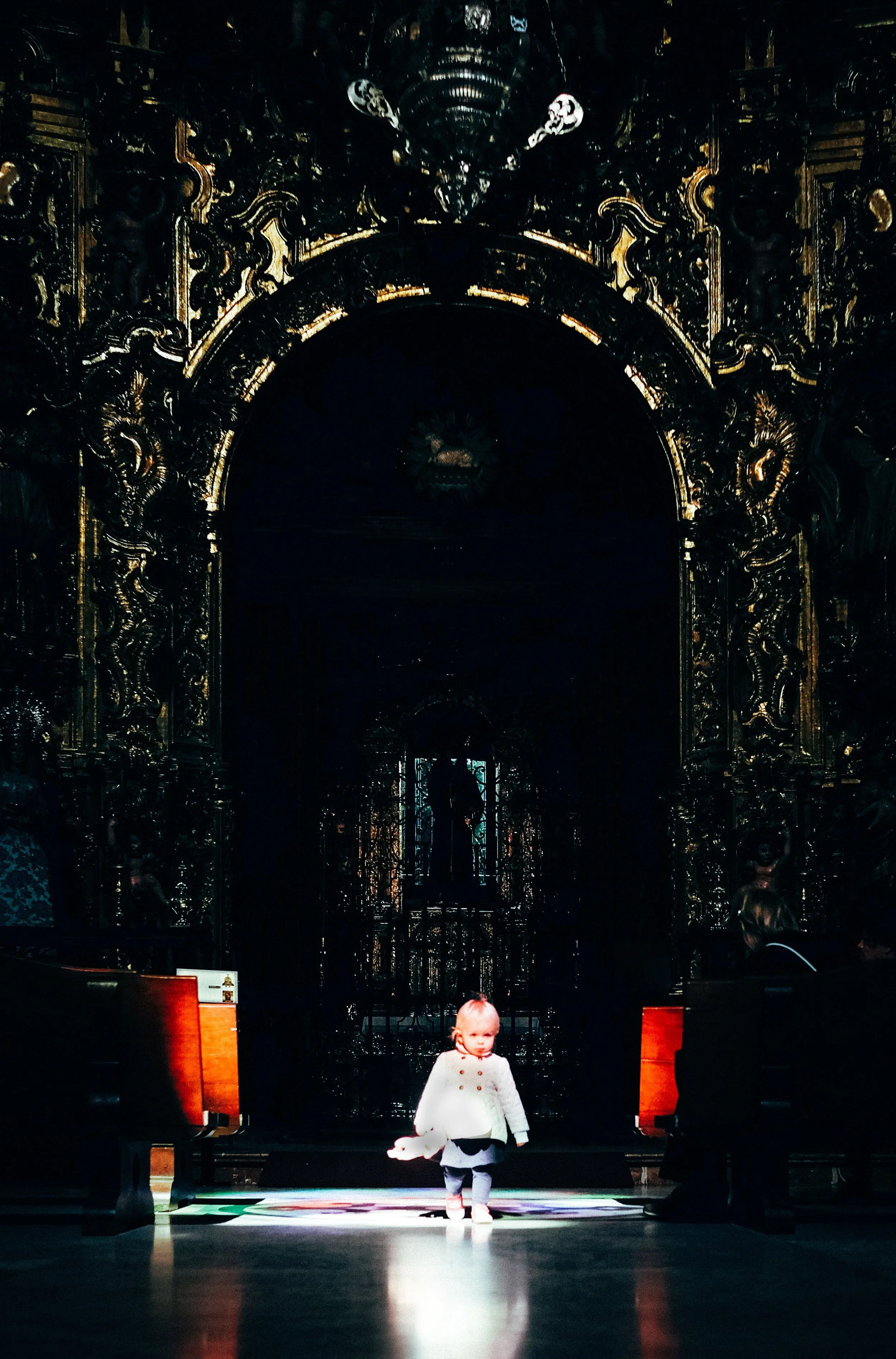A young girl walking down the aisle of a church or cathedral with ornate, dark wooden interior and intricate gold detailing. She is wearing a white coat and is brightly lit compared to the dim, richly decorated background.